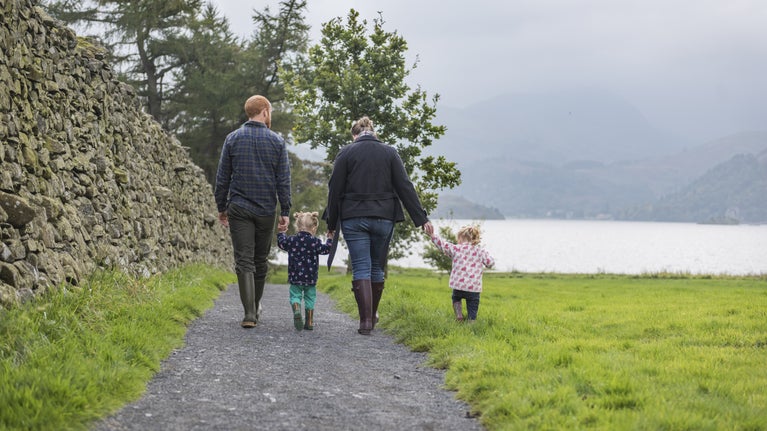 A family of four, two adults and two young children, hold hands as they walk down a path towards the lakeshore, against a mountain backdrop